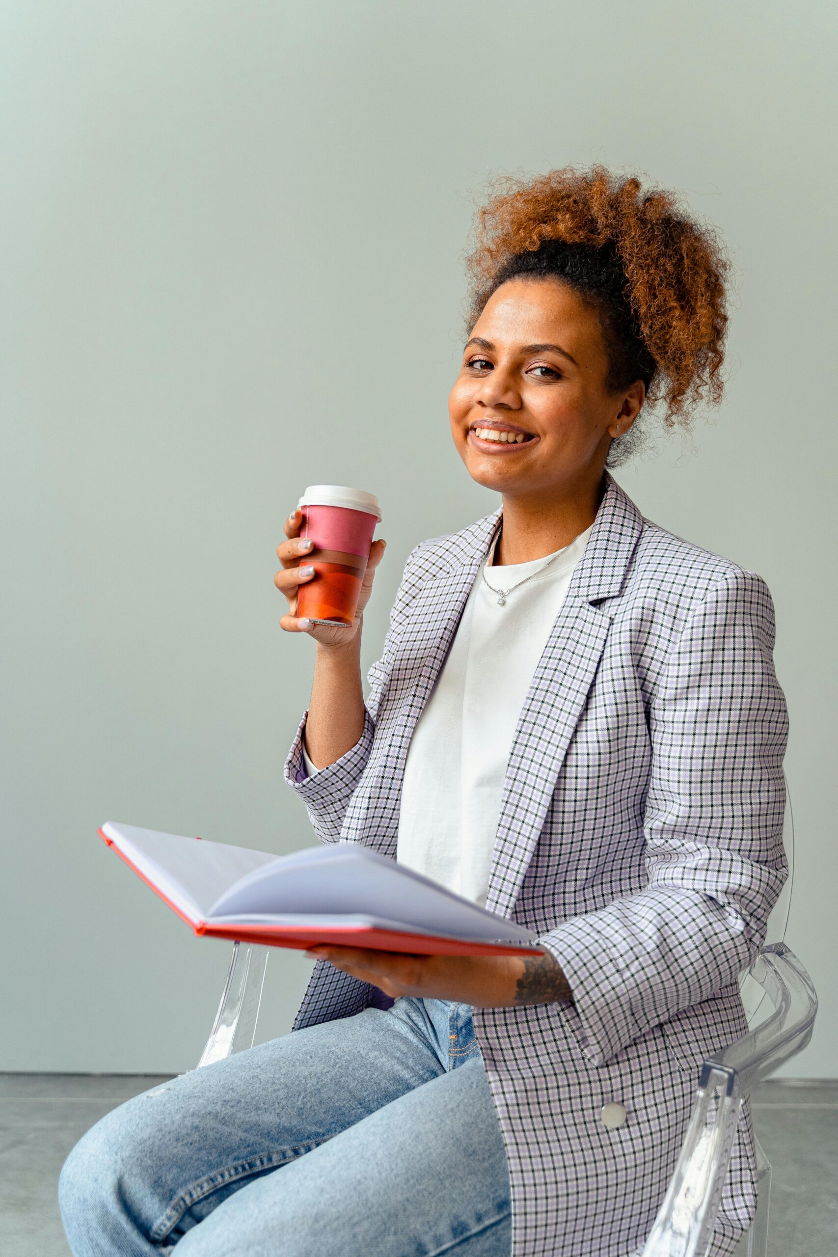 Smiling woman in blazer holds coffee cup and book, exuding confidence and warmth.